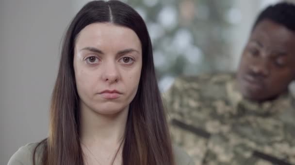 Hlavonosný portrét smutné, vystresované mladé ženy, dívající se do kamery jako rozmazaný voják křičící na pozadí. Detailní záběr zoufalé kavkazské mladé ženy a rozzlobený africký americký manžel. — Stock video