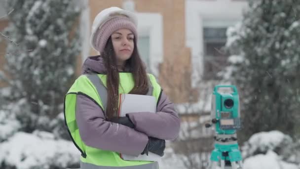 Középső lövés magabiztos pozitív női földmérő geodesist áll a szabadban nézi a kamera és mosolyog. Portré gyönyörű vékony professzionális kaukázusi nő pózol a szabadban hóesésben. — Stock videók