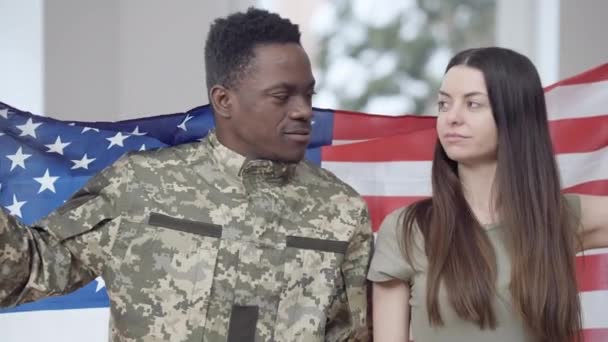 Homem afro-americano bonito feliz e mulher caucasiana posando com a bandeira dos EUA abraçando e sorrindo olhando para a câmera. Retrato de alegre casal inter-racial orgulhoso em casa. Conceito de patriotismo. — Vídeo de Stock