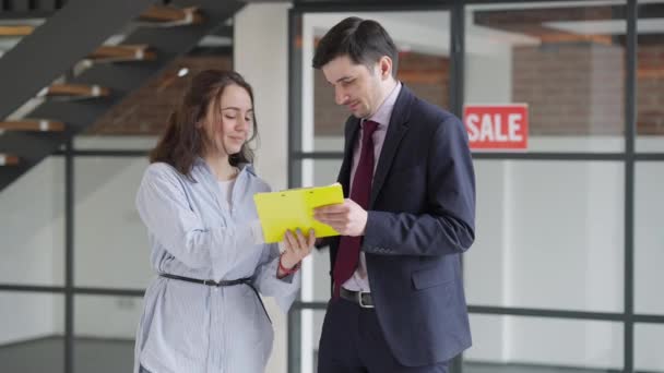 Cheerful young beautiful woman signing documents and receiving property key from man. Portrait of happy excited Caucasian flat or office owner with real estate agent indoors making agreement. — Stock Video