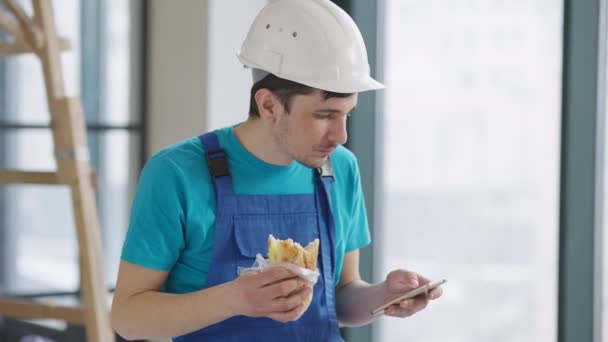 Meio tiro de reparador jovem construtor comer sanduíche e navegar na Internet no smartphone. Retrato do homem caucasiano a almoçar dentro de casa em novas instalações. Conceito de profissão. — Vídeo de Stock