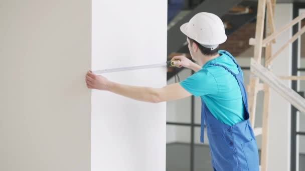 Vista lateral del hombre caucásico concentrado profesional en sombrero duro y pared de medición uniforme en interiores pensando. Retrato de constructor enfocado verificando locales. Concepto de industria. — Vídeos de Stock