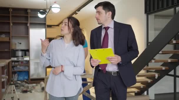 Joven corredor guapo caminando con el cliente en nuevas instalaciones como constructor que trabaja en el plan de proyecto de renovación en segundo plano. Retrato de hombre y mujer caucásicos confiados hablando examinando trabajos de reparación. — Vídeos de Stock