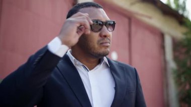 Kendine güvenen Afro-Amerikan bir adamın yakın çekimleri. Güneş gözlüklerini çıkarıyor. Etrafa bakıyor. Kasabada duran konsantre olmuş yakışıklı iş adamının portresi. Yaşam tarzı kavramı.