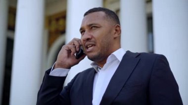 Afro-Amerikalı ciddi bir iş adamının dışarıda akıllı telefondan konuşmasının portresi. Takım elbiseli mutsuz yakışıklı adam telefonda bağırıyor. Stresli iş hayatı.