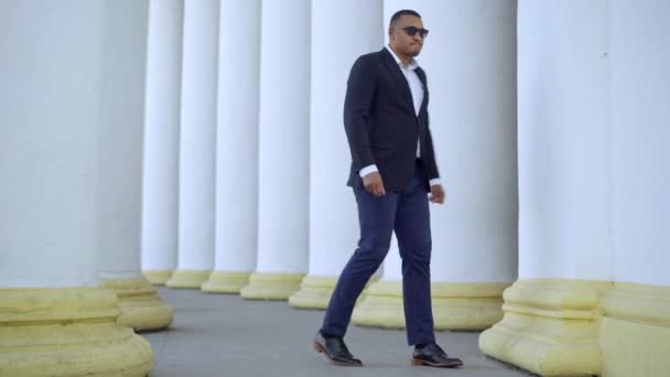 Wide shot of anxious worried African American man in formal suit and sunglasses walking at white columns. Portrait of stressed punctual businessman waiting for meeting outdoors in city. — Stock Video