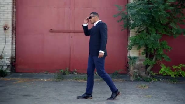 Vista lateral do homem afro-americano confiante em terno de negócios formal andando na favela urbana tirando óculos de sol. Retrato de tiro largo de empresário bem sucedido de volta em pequena cidade. — Vídeo de Stock