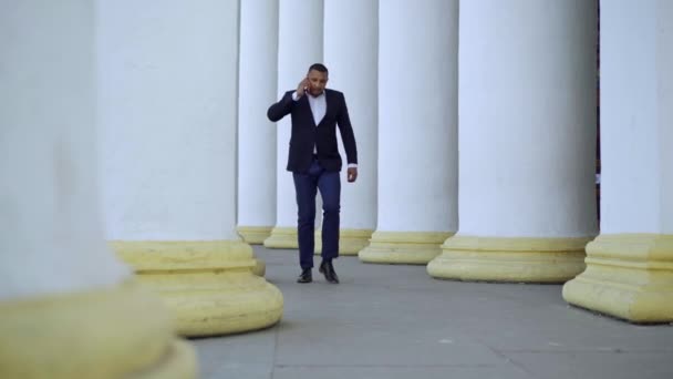 Amplio plano de ocupado hombre de negocios afroamericano hablando por teléfono caminando entre columnas blancas al aire libre y saliendo. Retrato de un hombre guapo serio y confiado discutiendo una idea de negocio. — Vídeos de Stock