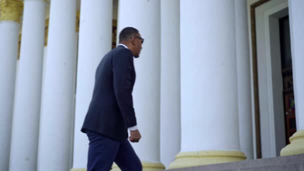 Sidovy av självsäker afroamerikansk affärsman i elegant kostym går uppför trappan utomhus med vita kolumner i bakgrunden. Lyckad man lämnar för affärsmöte. — Stockvideo