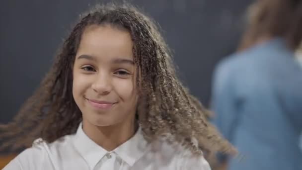 Portrait of charming confident smart African American schoolgirl with toothy smile posing in classroom with blurred classmate at background. Happy smiling girl looking at camera shaking head yes. — Stock Video