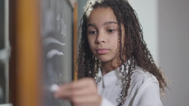 Colegiala zurda concentrada grave escribiendo con tiza en pizarra en el aula. Retrato de una hermosa chica afroamericana inteligente que estudia en la escuela en el interior. Enseñanza secundaria. — Vídeos de Stock