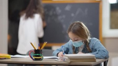 Koronavirüs maskeli beyaz kız öğrencinin sınıftaki masada yazı yazması ve başparmağını göstermesi. Arka plandaki tahtaya yazı yazan bulanık Afrikalı Amerikalı sınıf arkadaşı. Kapalı.