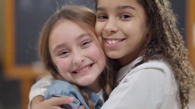 Kameraya bakıp sarılan ve gülümseyen neşeli liseli kızların yakın çekimi. Orta okulda sınıfta poz veren Afro-Amerikan ve beyaz kızlar. Arkadaşlık kavramı.