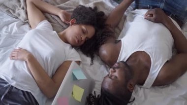 Tükenmiş Afro-Amerikan çift dizüstü bilgisayarla yatakta uyuyor. Yorgun, yakışıklı bir adam ve güzel bir kadının yatak odasında yatıp Covid salgınında aşırı çalışmasının en iyi görüntüsü..