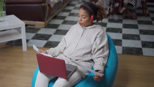 Znudzona Afroamerykanka w słuchawkach wystaje z języka analizując strategię na wykresach i wpisując na klawiaturze laptopa. Wysoki kąt widzenia wyczerpany młoda dama pracuje online w biurze domu. — Wideo stockowe