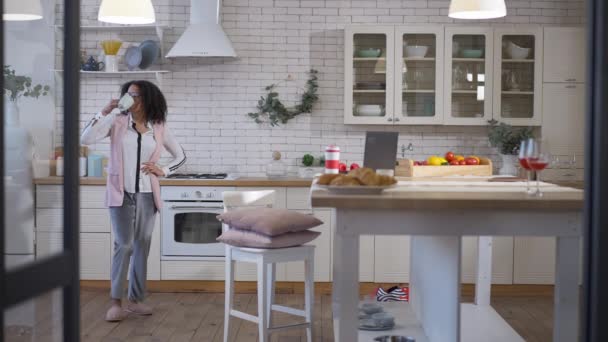 Amplio tiro de joven afroamericana mujer seria beber café de la mañana o té en la cocina y caminar a la mensajería portátil en línea. Retrato del exitoso freelancer en casa. — Vídeo de stock