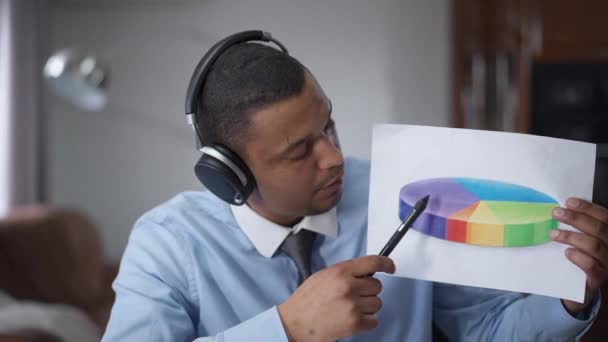 Professional African American analyst in headphones pointing at round diagram talking. Video chat POV of confident concentrated young man explaning business idea in virtual conference in home office. — Stock Video