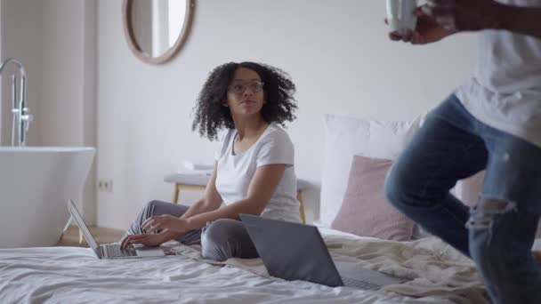 Gözlüklü Afro-Amerikan güzel bir eşin dizüstü bilgisayarda klavyeyle yazı yazması yatakta oturup, sevgi dolu yakışıklı kocayla kahve getirmesi. Bağımsız mutlu çiftler.. — Stok video