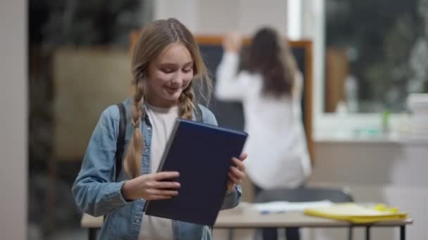 Charming smart Caucasian schoolgirl closing workbook and smiling looking at camera. Blurred African American classmate at background in classroom. Portrait of happy girl posing in school indoors. Stock Footage