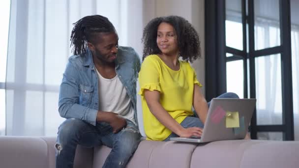 Amante jovem marido afro-americano abraçando esposa confiante trabalhando on-line a partir de casa. Positivo casal feliz olhando para a câmera sorrindo sentado no sofá na sala de estar. — Vídeo de Stock