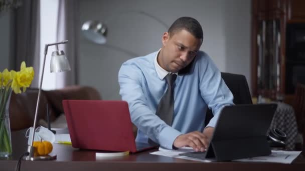 Μέση εικόνα του πολυάσχολου νεαρού Αφροαμερικανού επιχειρηματία να κλείνει τηλεφωνικά μηνύματα στο λάπτοπ και το tablet και να αναλύει γραφήματα. Πορτρέτο του σοβαρού επιτυχημένου άνδρα που εργάζεται online στο γραφείο στο σπίτι. — Αρχείο Βίντεο