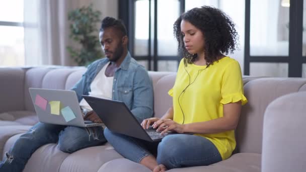 Enfocado delgado hermosa mujer afroamericana en los auriculares escribiendo en el teclado del ordenador portátil con la mensajería masculina guapo concentrado en línea en segundo plano. Pareja joven de freelancers en salón interior. — Vídeo de stock