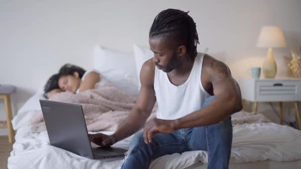 Homem Africano Americano concentrado bonito confiante digitando no teclado do laptop com a jovem mulher bonita dormindo na cama no fundo. Retrato de freelancer no escritório em casa dentro de casa. — Vídeo de Stock