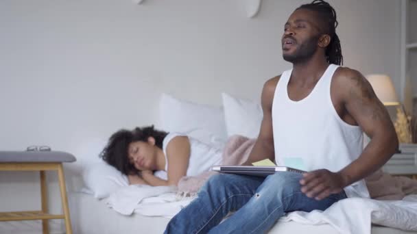 Portrait d'homme afro-américain tatoué satisfait assis sur le lit avec un ordinateur portable regardant femme endormie et pensant. Beau freelance confiant dans la chambre le matin ou le soir. Bureau à domicile. — Video
