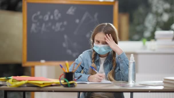 Portrait of overwhelmed junior high schoolgirl in Covid face mask holding head in hands sighing. Cute Caucasian girl sitting at desk in classroom looking at camera and shaking head. Pandemic concept. — Stock Video