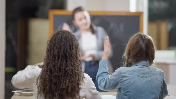 Estudantes do ensino médio sentados na mesa levantando a mão e fazendo perguntas desfocadas do professor. Visão traseira de colegas brancos e afro-americanos inteligentes diligentes que estudam no ensino fundamental. Educação. — Vídeo de Stock