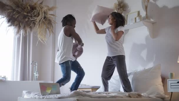 Ampla foto de alegre casal afro-americano lutando travesseiros no quarto com laptop na cama. Jovem positivo e mulher tendo pausa trabalhando on-line em casa no isolamento pandêmico. — Vídeo de Stock