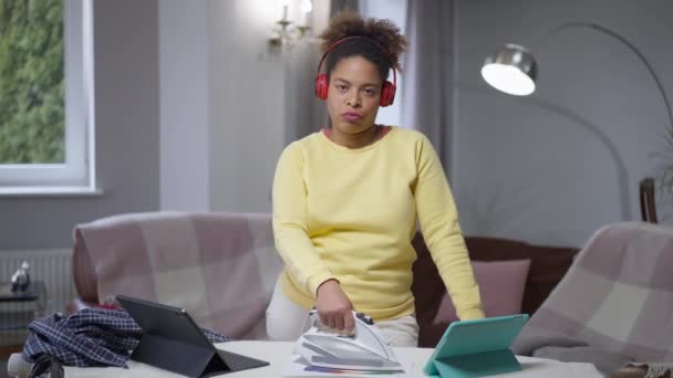 Overbelaste vermoeide Afro-Amerikaanse vrouw strijken diagram staan in het thuiskantoor kijken naar de camera met uitgeputte ontevreden gezichtsuitdrukking. Portret van overwerkte dame op coronavirus lockdown. — Stockvideo