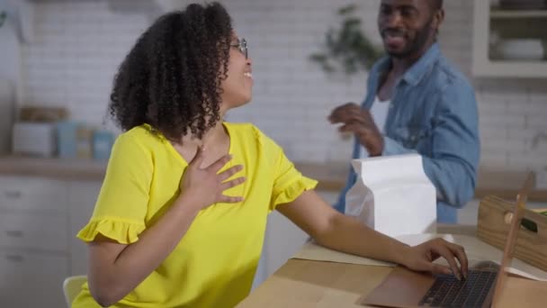 Tacksam vacker afroamerikansk kvinna ler mot mannen med mat leverans. Porträtt av överarbetad ung fru messaging online på laptop sitter i köket hemma. Ny normal livsstil. — Stockvideo