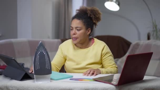 不安は、アフリカ系アメリカ人の女性がノートパソコンやタブレットにキーボードを入力し、自宅のオフィスでアイロン台に座って強調した。屋内でオンラインで働く圧倒的な若いマネージャーの肖像画. — ストック動画