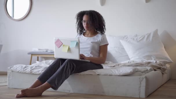 Amplio tiro de hermosa exitosa mujer afroamericana mensajería en línea escribiendo en el teclado del ordenador portátil y cayendo de nuevo en la cama. Retrato de freelancer cansado y sobrecargado de trabajo trabajando en casa en interiores. — Vídeos de Stock