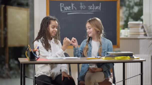 Pinky jura sorrir lindas alunas na sala de aula. Feliz alegre meninas caucasianas e afro-americanas tremendo dedos pequenos e abraçando sentado na escola dentro de casa. Conceito de amizade e confiança. — Vídeo de Stock