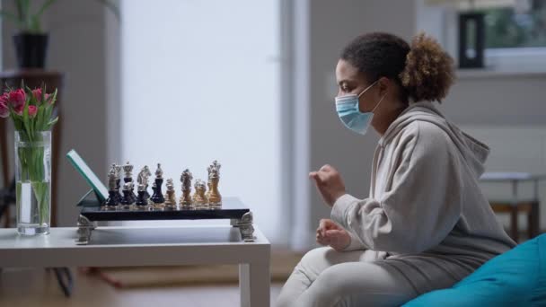 Side view of young African American intelligent woman in coronavirus face mask playing chess in front of tablet with live streaming. Confident smart lady on Covid-19 isolation at home indoors. — Stock Video