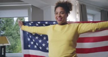 Amerikan bayrağına sarılmış, gülümseyen Afro-Amerikan genç bir kadın. Memleketinde ulusal sembolle poz veren kendinden emin mutlu bir kadının portresi. Sinema 4k ProRes Merkezi.