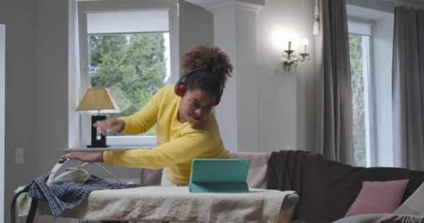 Pozitivní mladá Afroameričanka žehlení prádla s jednou rukou a mluvení na tablet webkamery s úsměvem. Portrét přepracovaného nezávislého pracovníka v domácí kanceláři na pandemii koronaviru. Cinema 4k ProRes HQ — Stock video