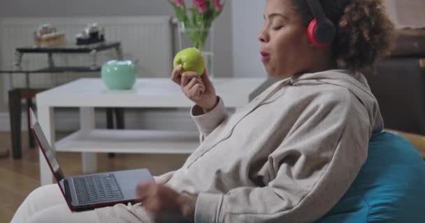 Vue latérale de la jeune femme dans les écouteurs mordant mâcher des messages de pomme saine en ligne sur ordinateur portable. Gestionnaire afro-américain confiant assis sur une chaise longue dans son bureau. Siège social Cinema 4k ProRes. — Video