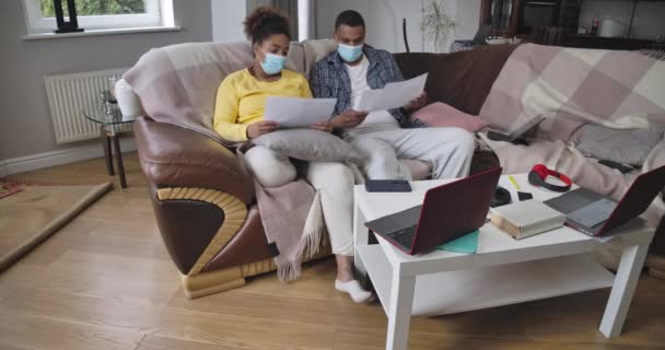 Wide shot portrait of African American man and woman in coronavirus face masks sitting on couch analyzing graphs talking. Smart young husband and wife in home office. Cinema 4k ProRes HQ. — Stock Video