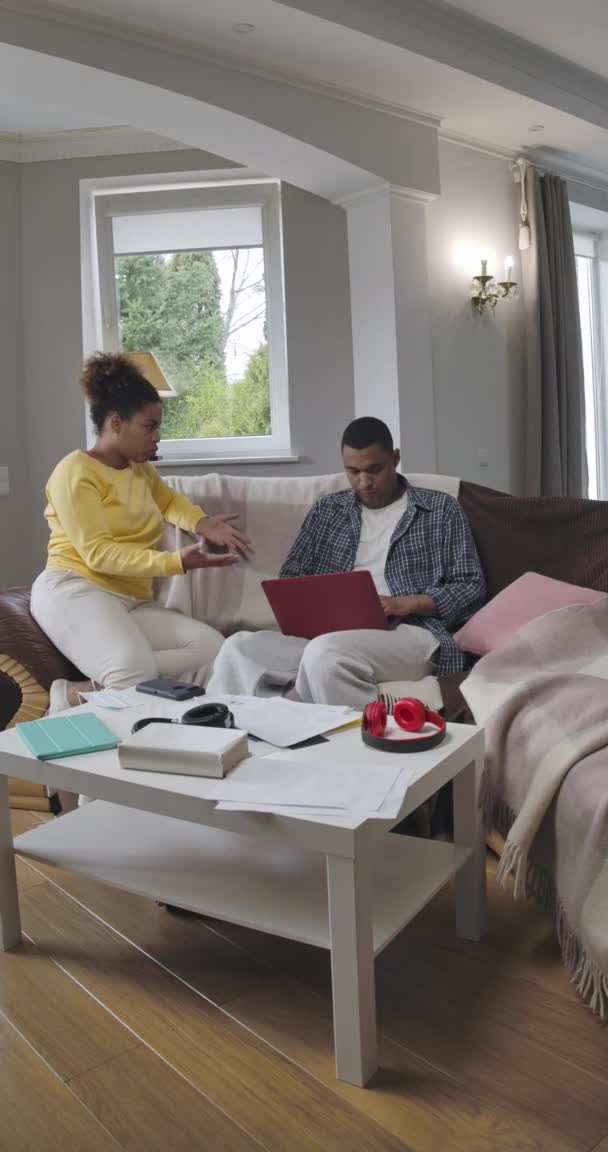 Vídeo vertical da mulher afro-americana irritada irritada discutindo com o homem ocupado digitando no teclado do laptop. Absorvido jovem marido ignorando esposa trabalhando on-line no escritório em casa. Cinema 4k ProRes HQ. — Vídeo de Stock