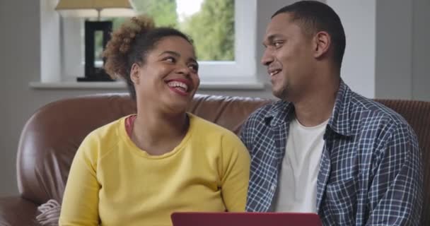 Gelukkig stel dat thuis zit te glimlachen en knuffelen. Positief liefdevolle Afro-Amerikaanse man en vrouw genieten van werken op afstand zittend op de bank in de woonkamer. Cinema 4k ProRes Hoofdkwartier. — Stockvideo