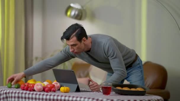 Jovem focado homem do Oriente Médio assistindo receita on-line em tablet de pé à mesa com frutas e legumes. Chef bonito concentrado com ingredientes crus na cozinha em casa. — Vídeo de Stock