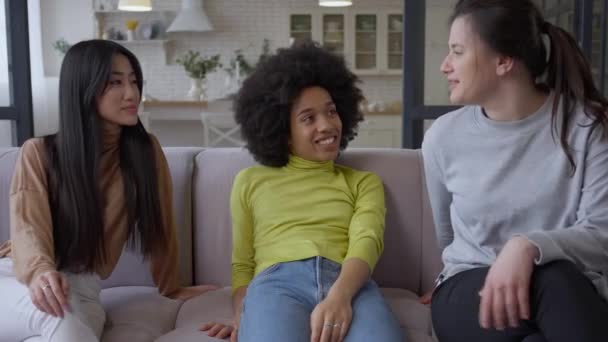 Vrolijke jonge multinationale vrouwen die op de bank zitten te praten. Positieve blanke Aziatische en Afro-Amerikaanse vrouwelijke vrienden roddelen binnen in de woonkamer. Vriendschap en vrije tijd. — Stockvideo