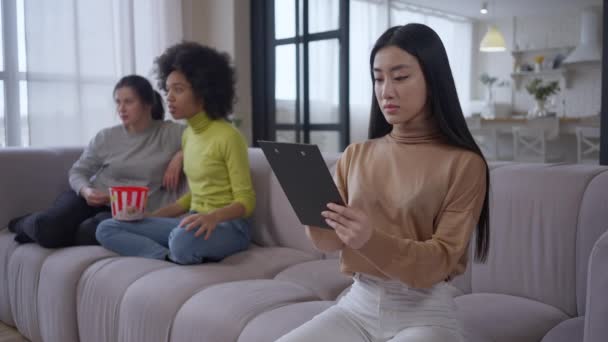 Selbstbewusste, ernsthafte Asiatin, die sich Dokumente ansieht, während sie im Homeoffice auf der Couch sitzt und im Hintergrund multiethnische Mitbewohner fernsehen. Konzentrierte Smart Lady auf Fernarbeit. — Stockvideo