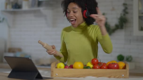 Gledelig ung afrikansk kvinne med hodetelefoner som danser med kjevle i hånden. Varsom, lystig, vakker dame som har det gøy på kjøkkenet mens hun lager sunn middag. Livsstilskonsept. – stockvideo