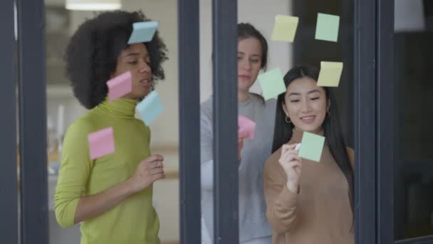 Plan médian de trois jeunes femmes multiethniques concentrées collant des notes sur le miroir parlant. De belles femmes multinationales confiantes qui travaillent au bureau à domicile pour discuter d'une idée de start-up. Concept de travail d'équipe. — Video