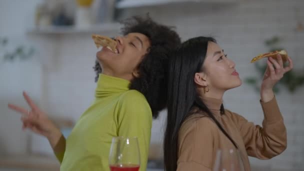 Funny young African American and Asian women standing back to back in kitchen eating pizza. Positive happy carefree friends having fun indoors. Friendship and joy concept. — Stock Video