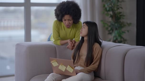 Sad young Asian woman sitting on couch with laptop as African American friend calming down supporting lady. Portrait of upset beautiful women in home office working online. Stress and support. — Stock Video